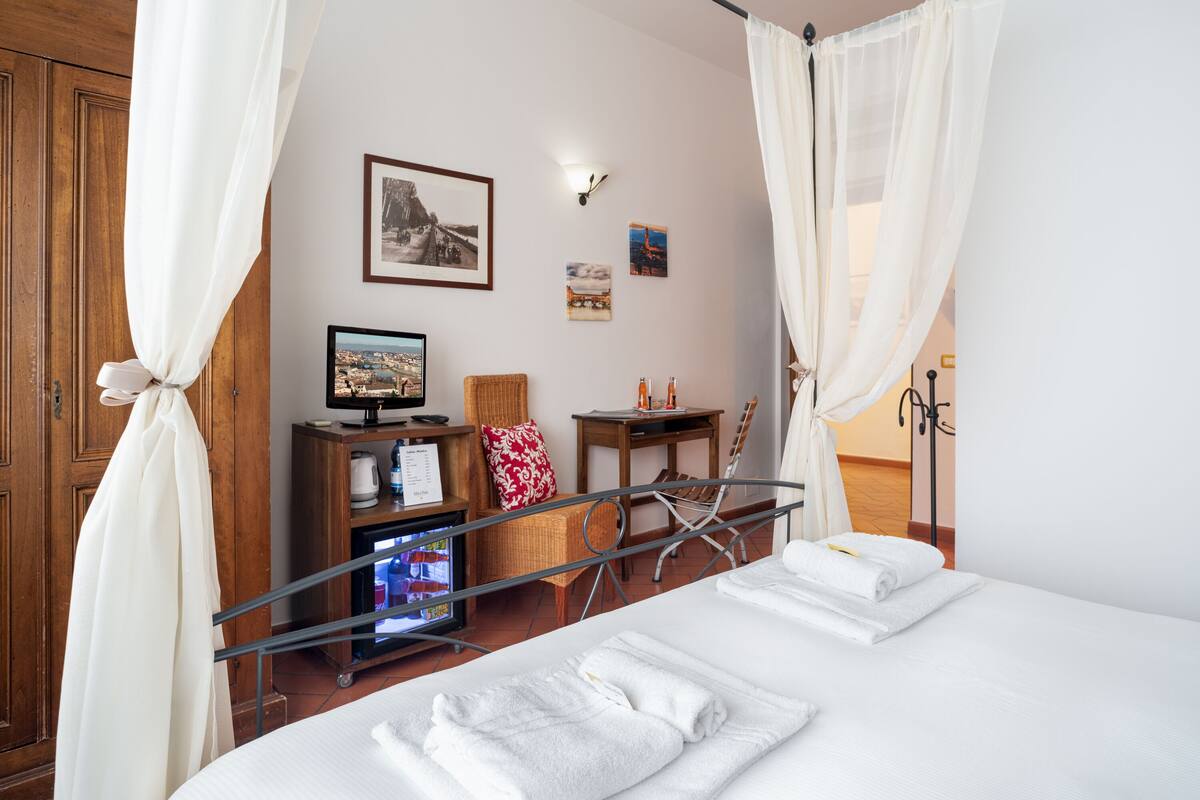 A hotel room with a bed, a desk with a computer, a chair, and a small table with drinks.