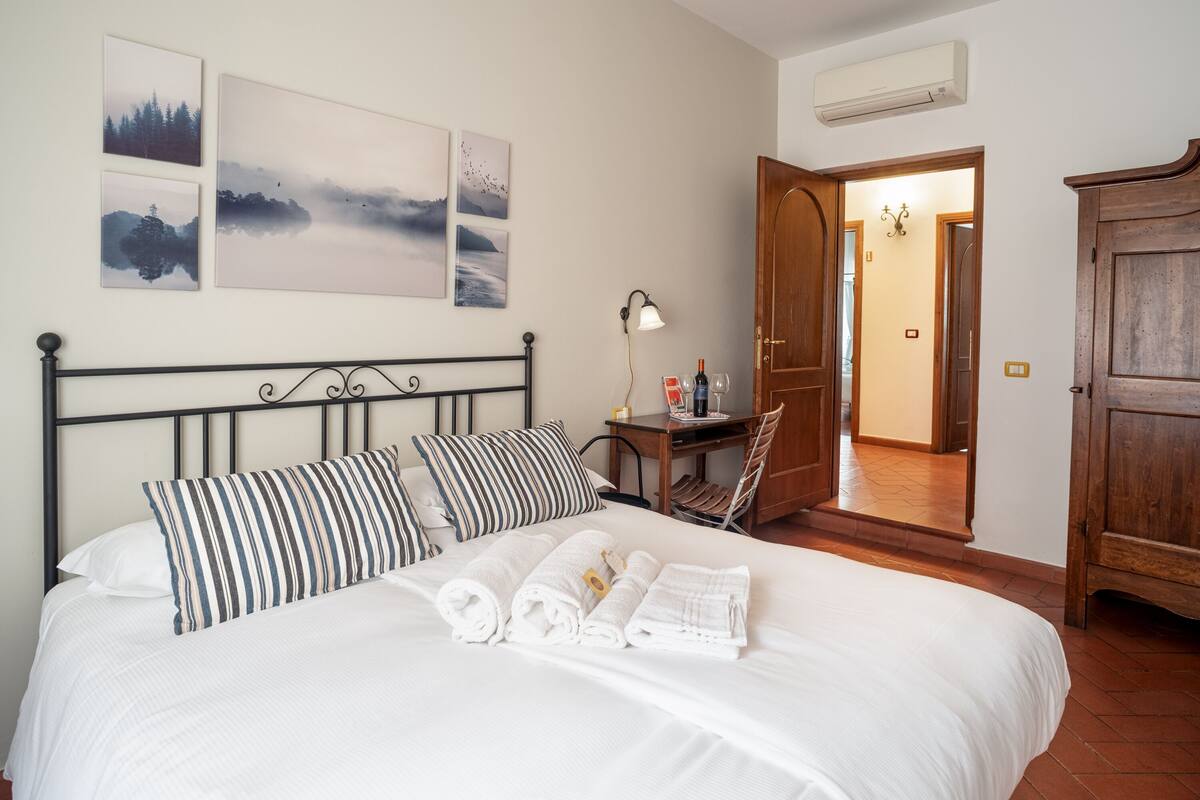 A neatly made bed with striped pillows, a bedside table with a lamp and a bottle, and artwork on the wall.