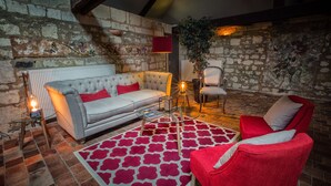 A cozy living room with a stone wall, a grey sofa with red cushions, a red armchair, a patterned rug, and a potted plant. - Le Chai de la Paleine (Le Puy-Notre-Dame)