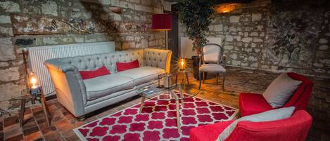 Una sala de estar acogedora con una pared de piedra, un sofá gris con cojines rojos, un sillón rojo, un tapiz estampado y una planta en maceta.