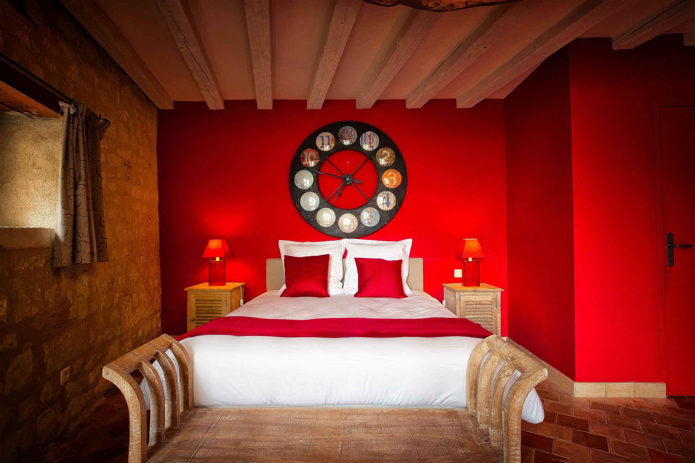 Une chambre avec un mur rouge, une horloge ronde, un lit avec une literie blanche et des oreillers rouges, deux tables de chevet avec des lampes et un banc en bois au pied du lit.