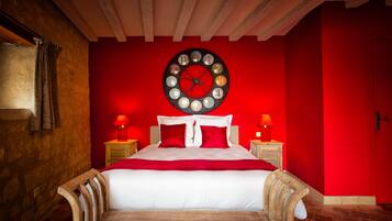 A bedroom with a red wall, a round clock, a bed with white bedding and red pillows, two bedside tables with lamps, and a wooden bench at the foot of the bed.