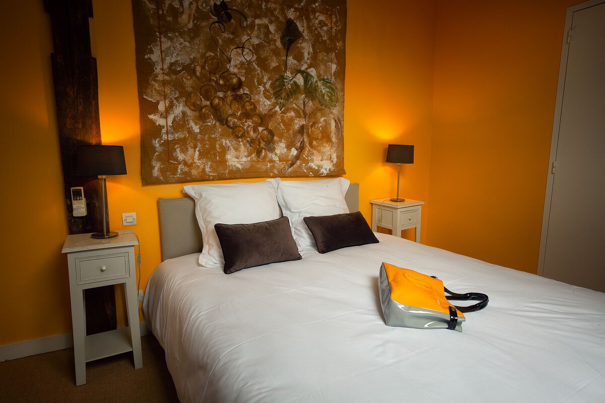 Una cama bien hecha con almohadas blancas y marrones, una mesita de noche con lámpara y una obra de arte en la pared sobre la cama.
