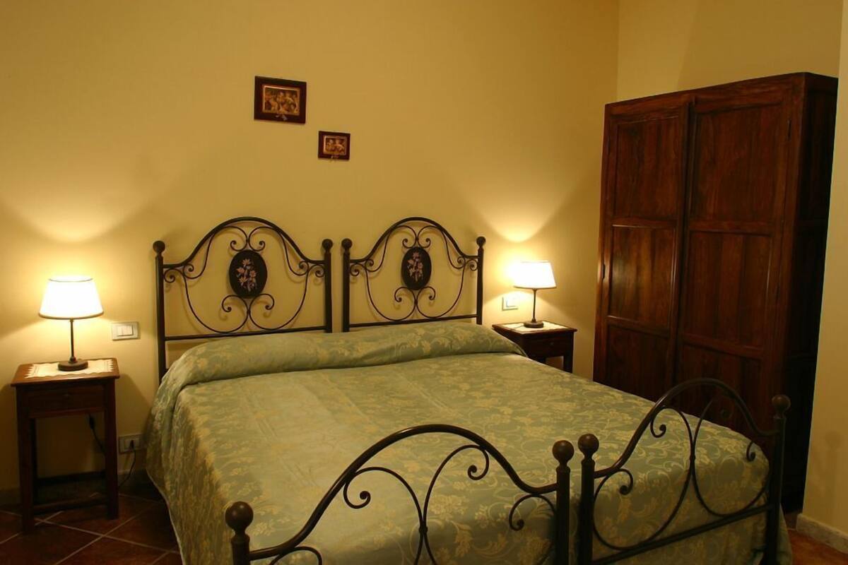 Un dormitorio con una cama, dos mesitas de noche con lámparas, un armario de madera y dos cuadros enmarcados en la pared.