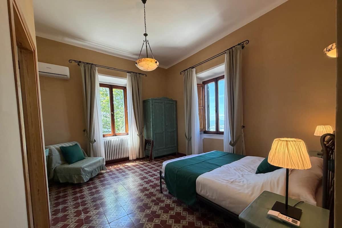 A hotel room with a bed, a chair, a desk with a lamp, a wardrobe, and two windows with curtains.