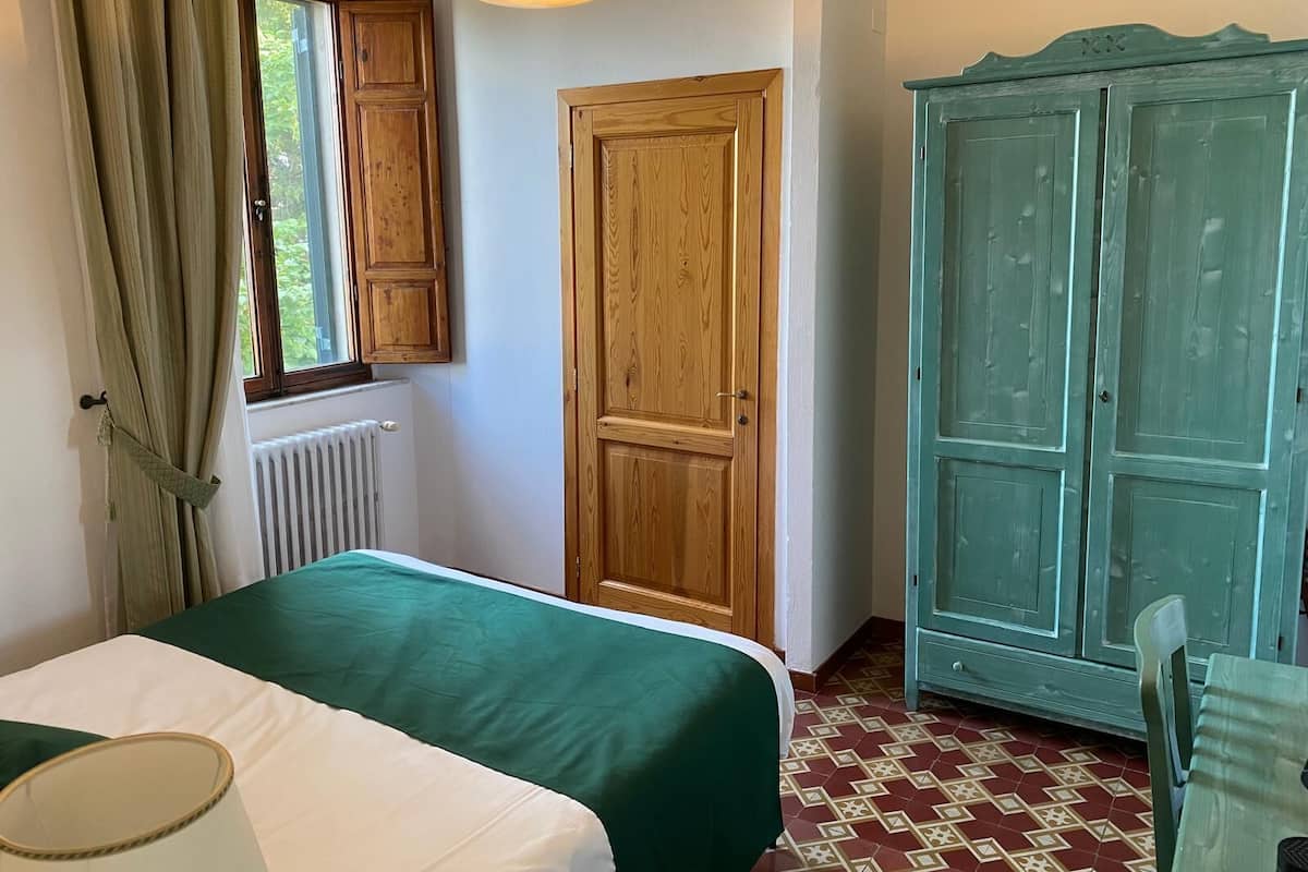 A room with a bed, a window with curtains, a radiator, a wooden door, and a green cabinet.