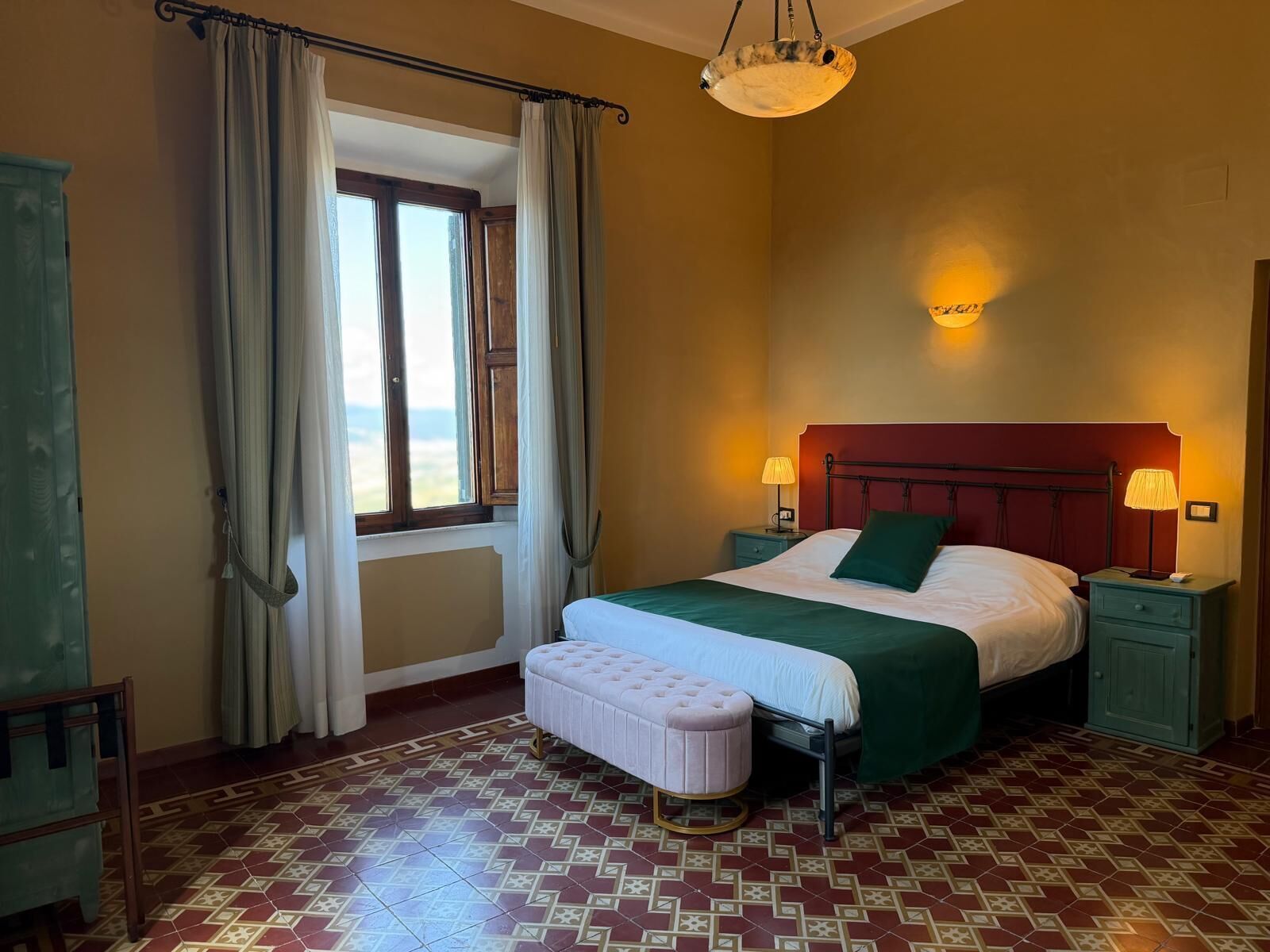 A bedroom with a bed, bedside lamps, a window with curtains, a bench, and patterned floor tiles.