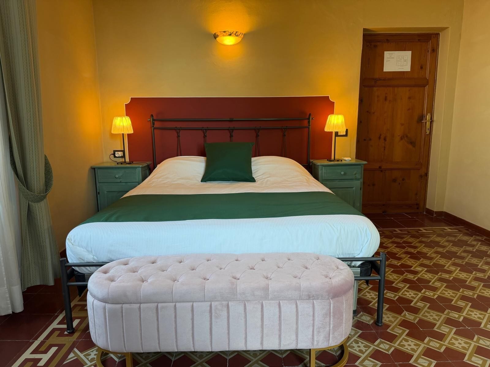 A neatly made bed with a green pillow, two bedside lamps, a wooden door, and a patterned floor.