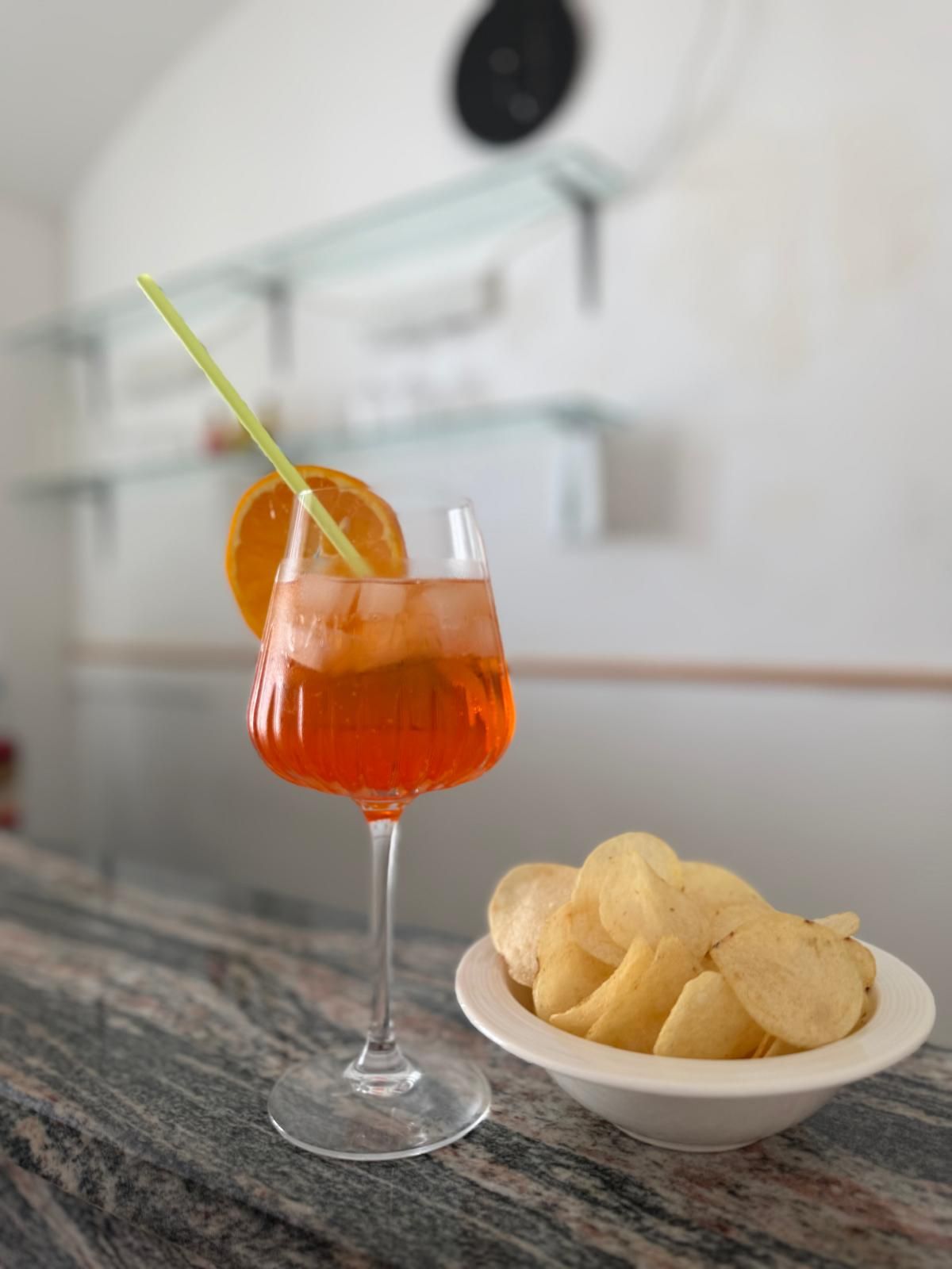 A cocktail glass with an orange slice garnish on a marble countertop, accompanied by a bowl of chips.