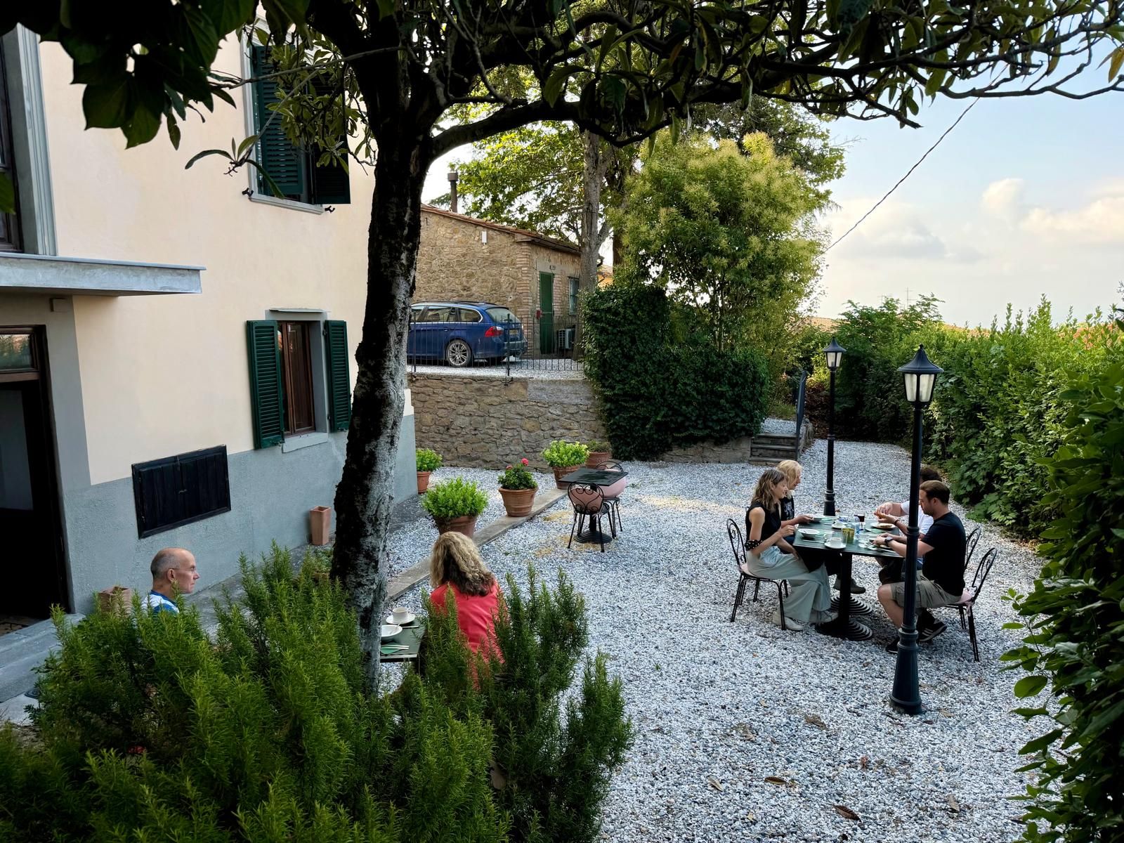 An outdoor patio with tables and chairs, surrounded by greenery and a building with stone walls.