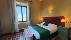 A bedroom with a bed, two bedside lamps, a window with curtains, a view of the landscape, and a patterned floor. - Villa Porta all'Arco (Volterra)