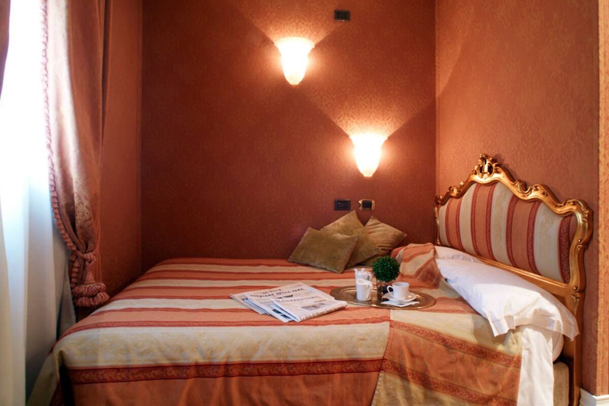 A hotel room with a large bed, two wall-mounted lights, a decorative headboard, and a small plant on the bedside table.