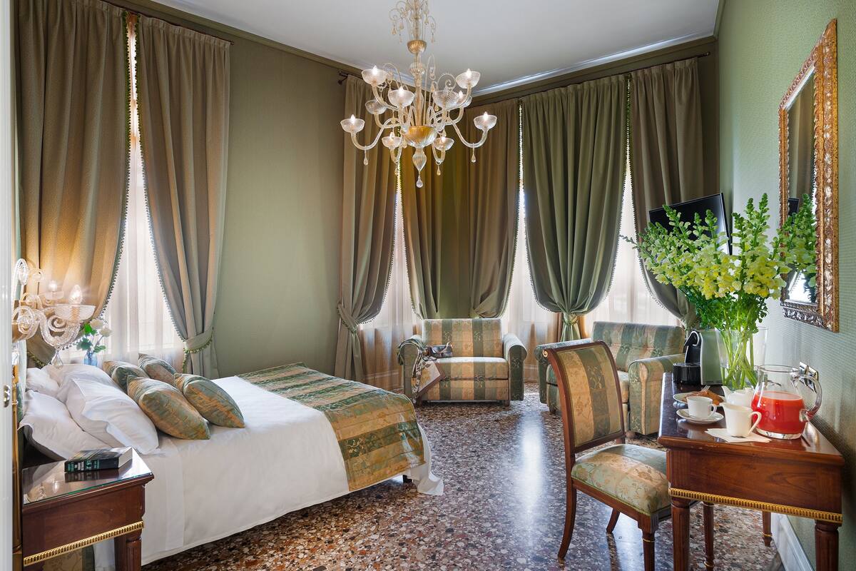 A hotel room with a bed, two armchairs, a wooden side table, a chandelier, a mirror, and a vase with flowers.