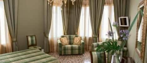 Una habitación con cama, sofá, sillón, una mesa con un florero y una araña.