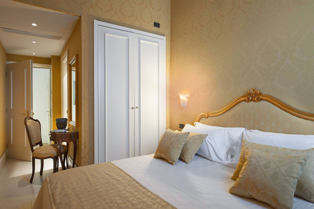A hotel room with a bed, two pillows, a nightstand, a chair, and a desk with a lamp.