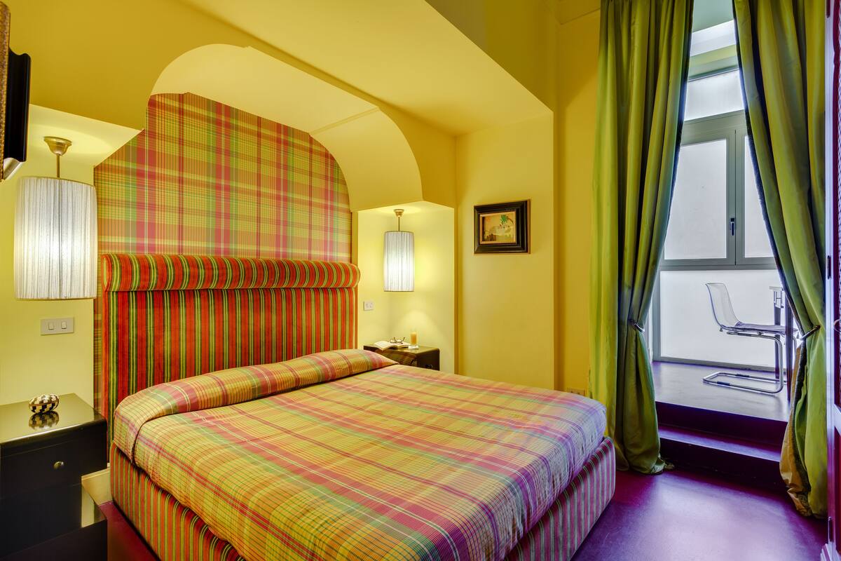 Habitación de hotel con cama, mesita de noche, lámpara, ventana con cortinas y vista al exterior.