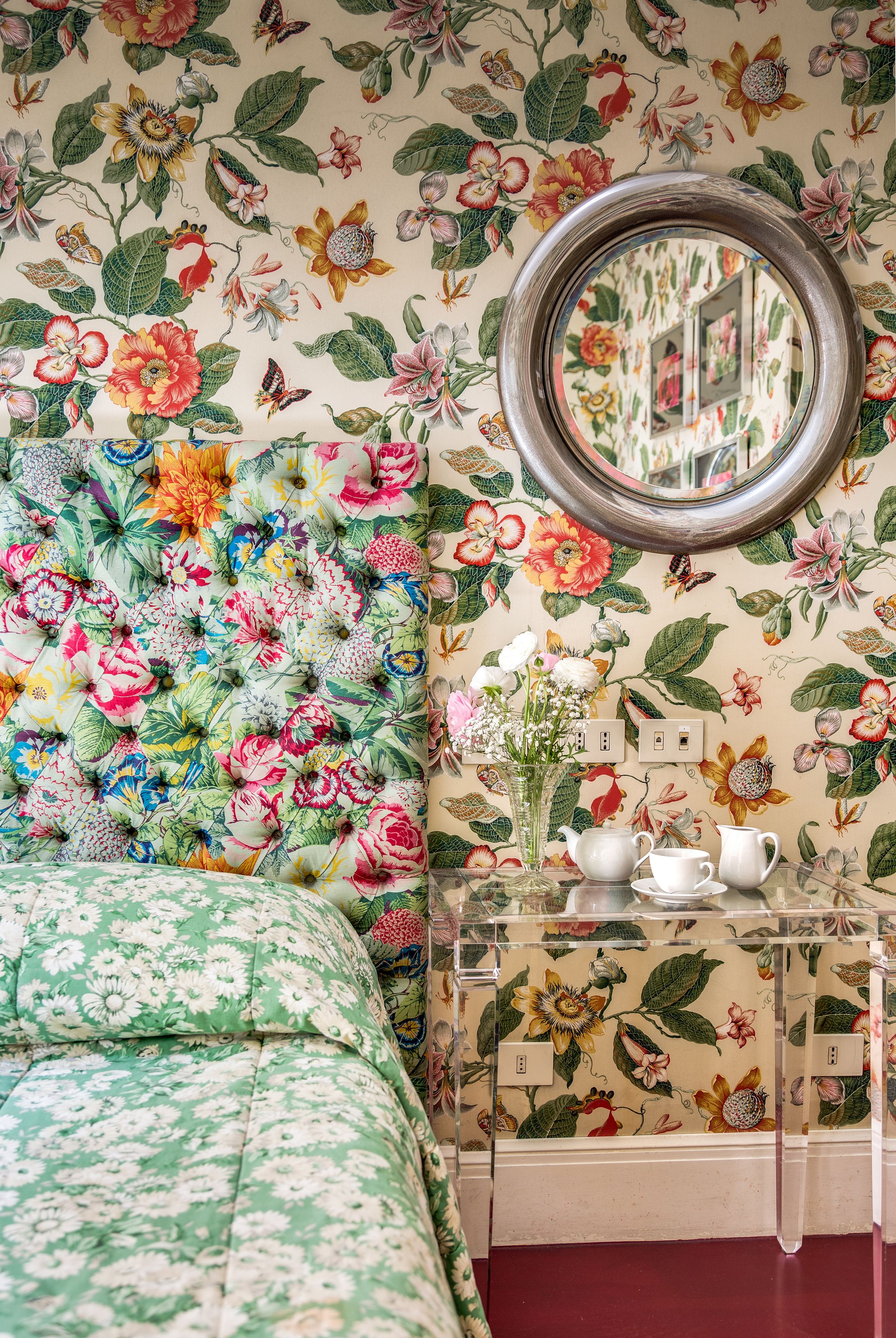 A room with floral wallpaper, a round mirror, a clear side table with a vase and teapots, and a green floral bedspread.
