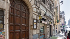 Un edificio histórico con una gran puerta de madera, un restaurante llamado "La Pizzeria" y una calle con coches estacionados y peatones.