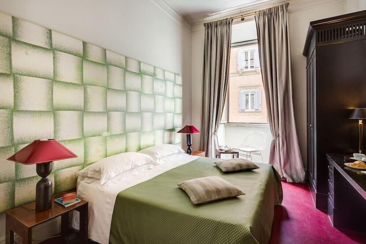 Habitación de hotel con una cama grande, una mesita de noche con lámpara, una silla, un escritorio con televisor y una ventana con cortinas.