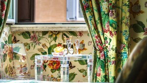 Una habitación con papel tapiz floral, una ventana con cortinas y una mesa de vidrio transparente con botellas y vasos.