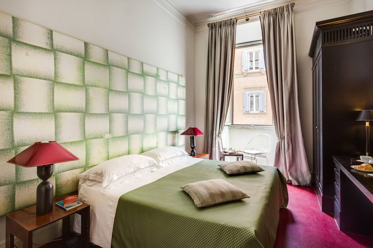 Habitación de hotel con una cama grande, una mesita de noche con lámpara, una silla, un escritorio con lámpara y vistas a un edificio a través de la ventana.