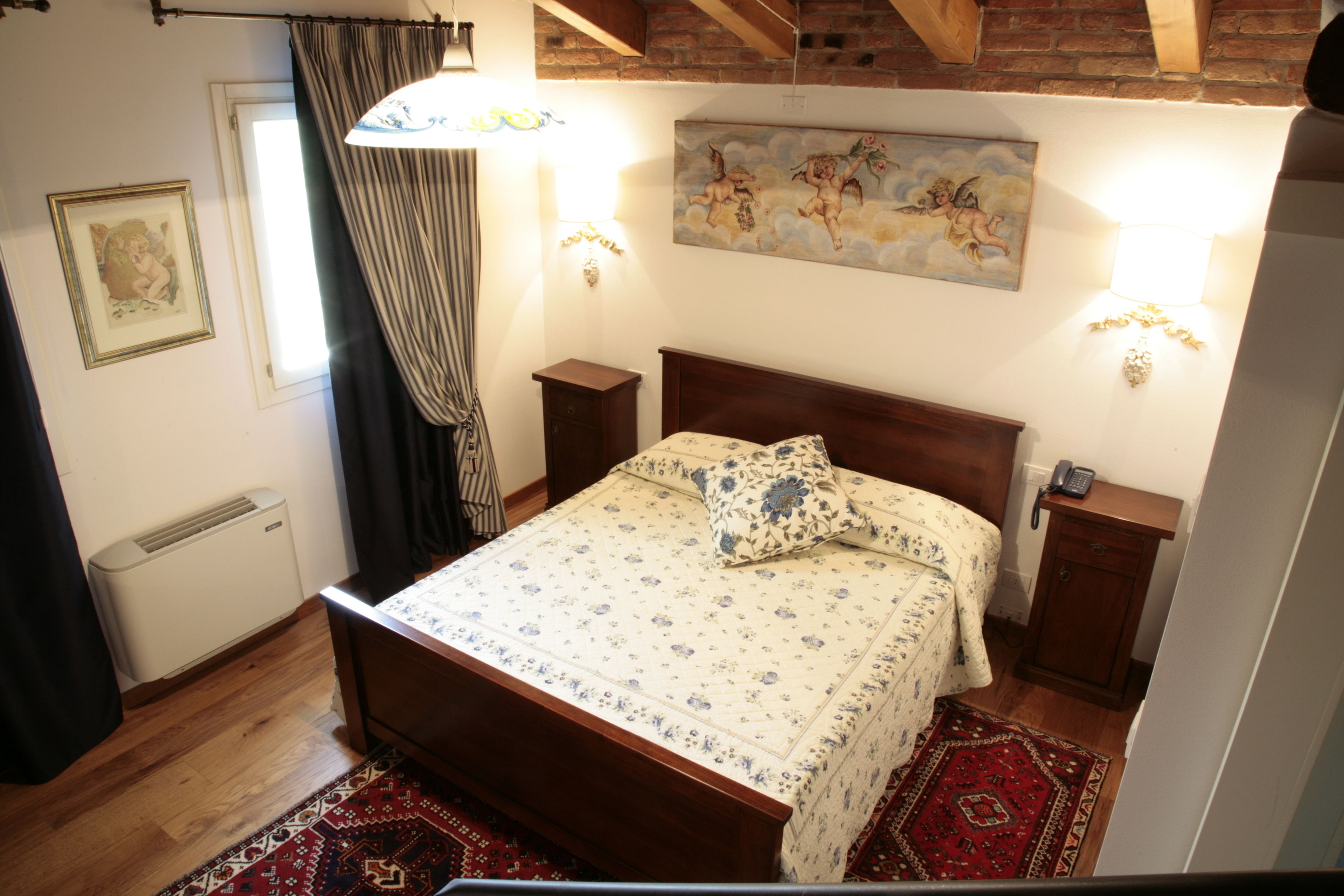 A bedroom with a wooden bed, floral bedding, a red patterned rug, a painting of cherubs on the wall, and a telephone on a nightstand.