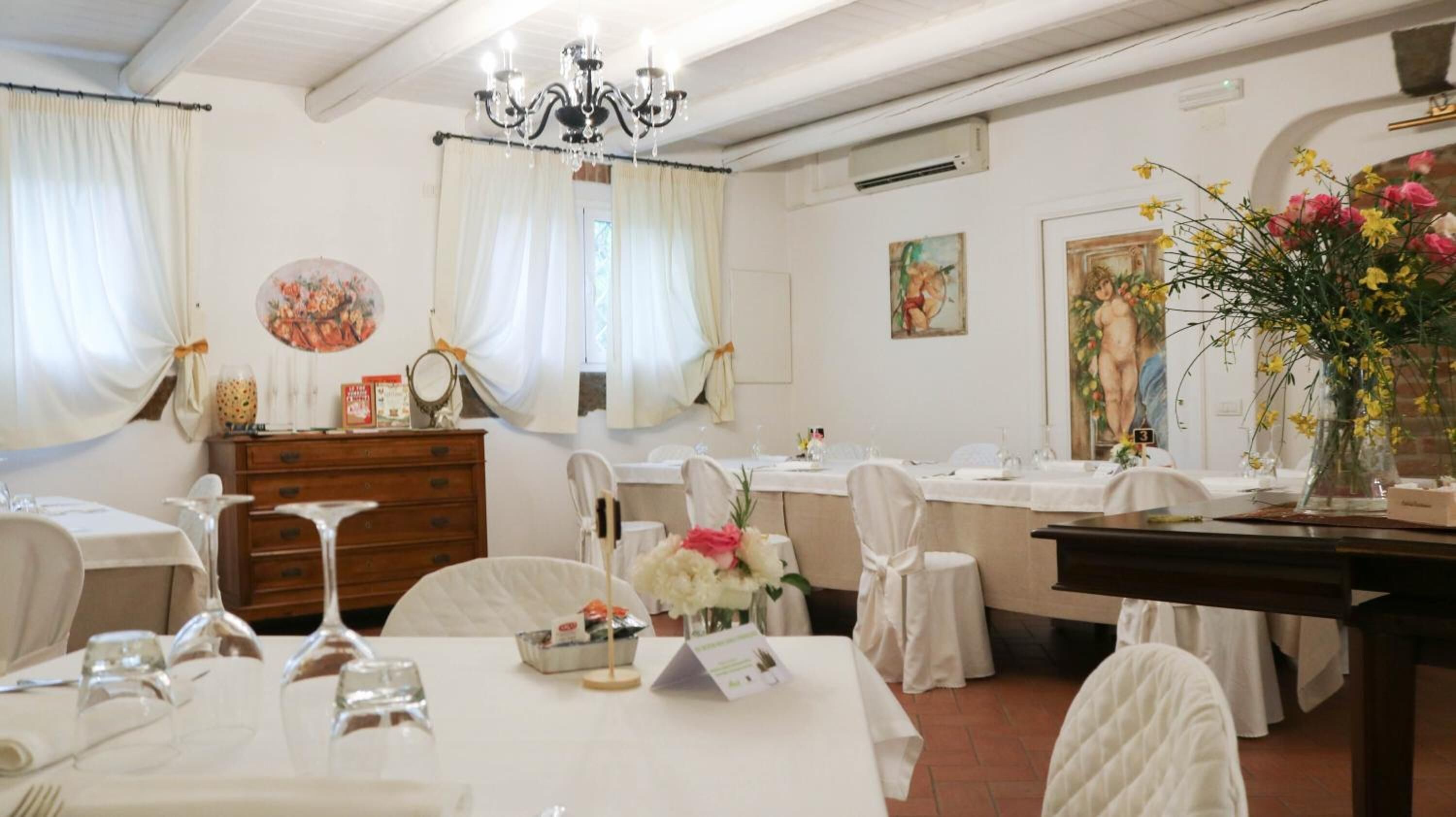 Une salle à manger avec des nappes blanches, des chaises et un lustre. Les murs sont ornés de tableaux et un vase rempli de fleurs est posé sur la table.