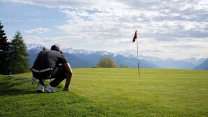 A person on a golf course with a flagstick, mountains in the background, and a clear sky. - Olympic (Crans-Montana)