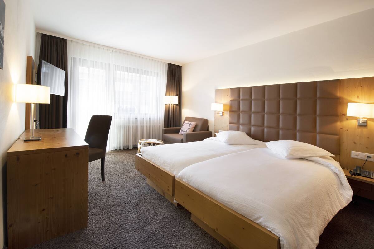 Quarto de hotel com duas camas, uma escrivaninha, uma cadeira, uma televisão e uma janela grande com cortinas.