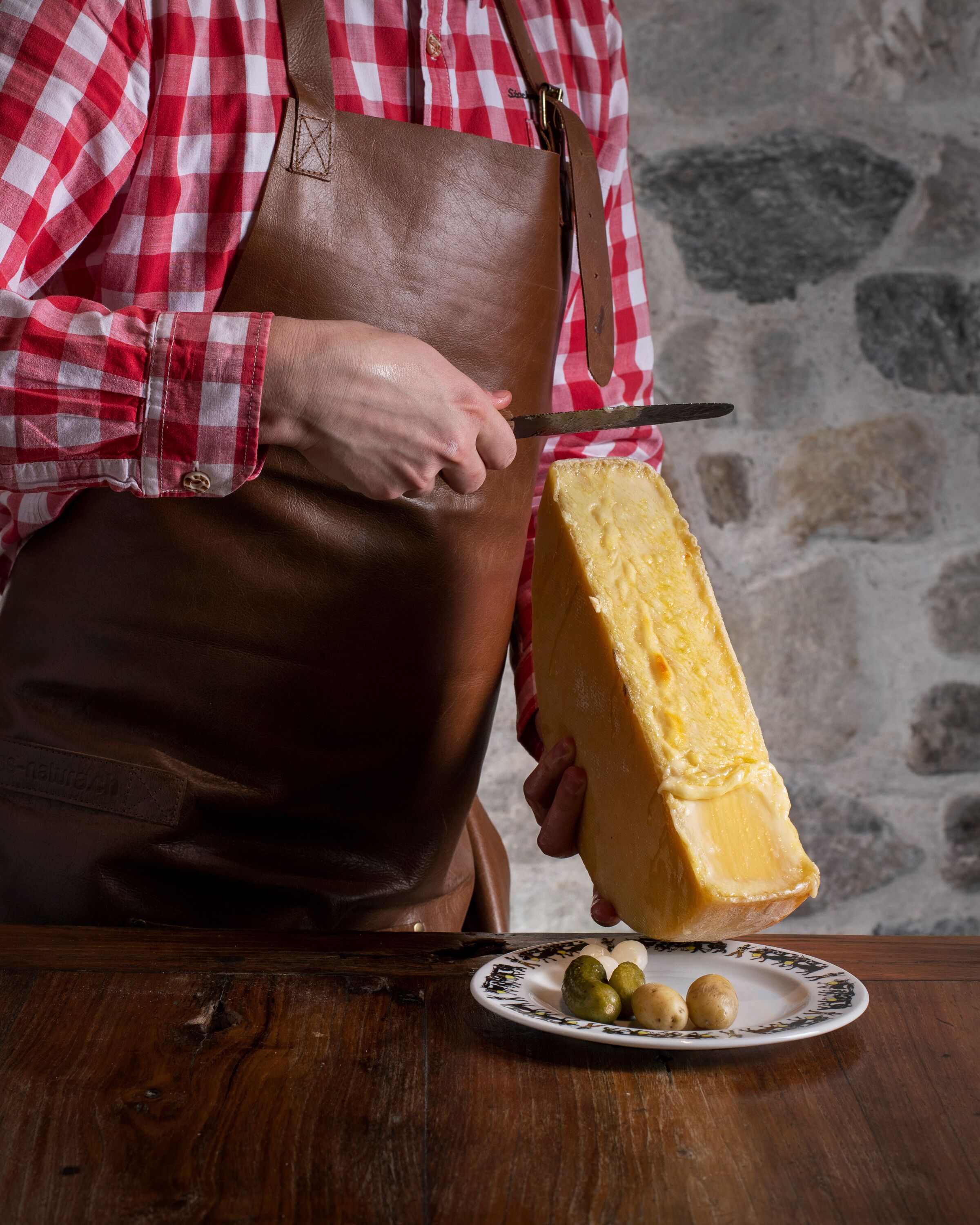 A person cutting a large block of cheese on a wooden table with a plate of olives nearby.