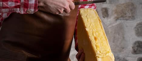 Une personne est en train de découper une grosse portion de fromage sur une table en bois, avec une assiette d’olives à proximité.