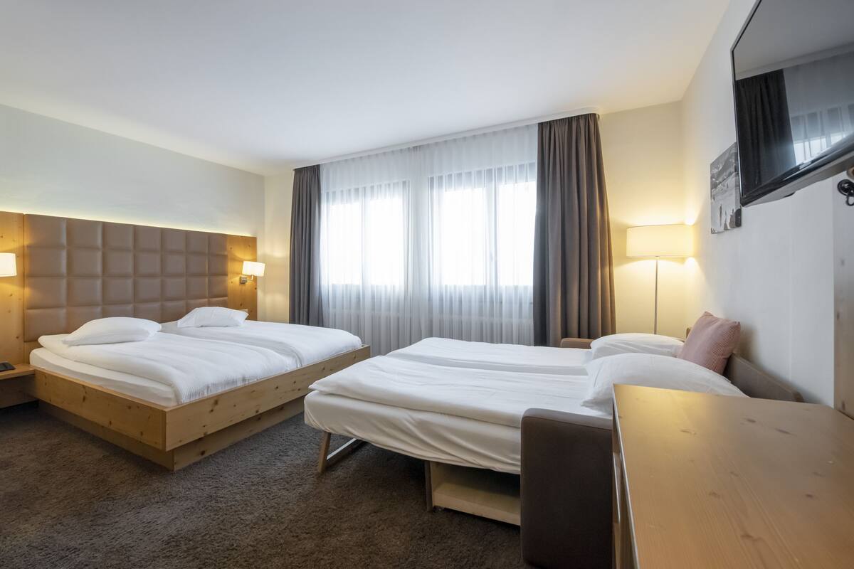 Quarto de hotel com duas camas, uma escrivaninha de madeira e uma janela grande com cortinas.