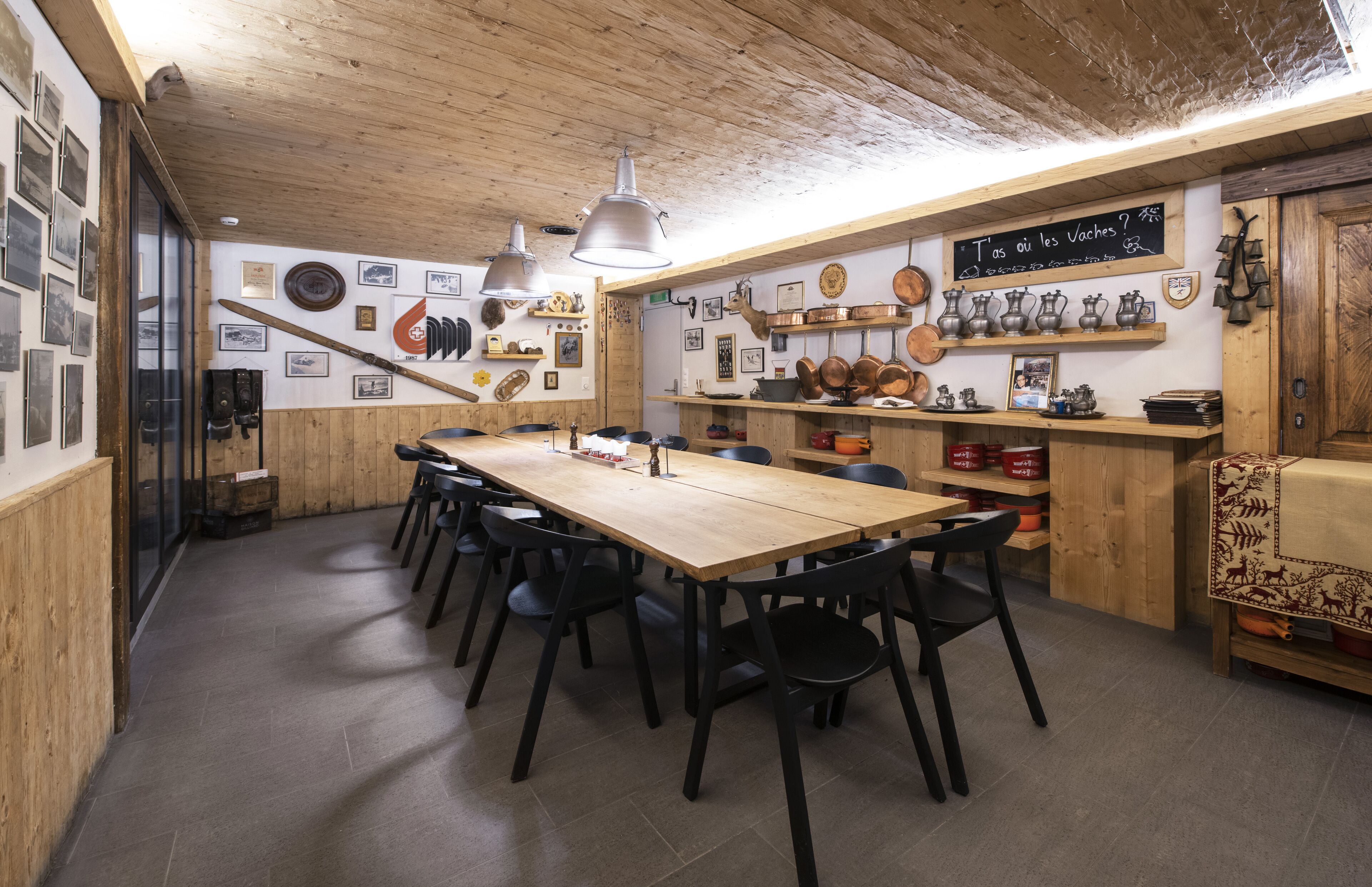 A rustic dining area with a long wooden table, black chairs, and a variety of kitchen utensils displayed on the walls.