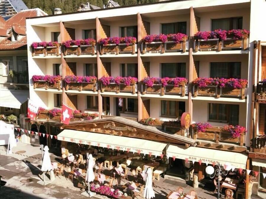 A multi-story hotel with balconies adorned with flowers, a ground-level bar, and a mountainous backdrop.