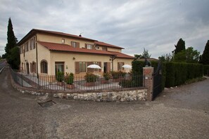 A two-story house with a tiled roof, a stone foundation, and a fenced patio area with potted plants and umbrellas. - Casa Santa Caterina (Asciano)