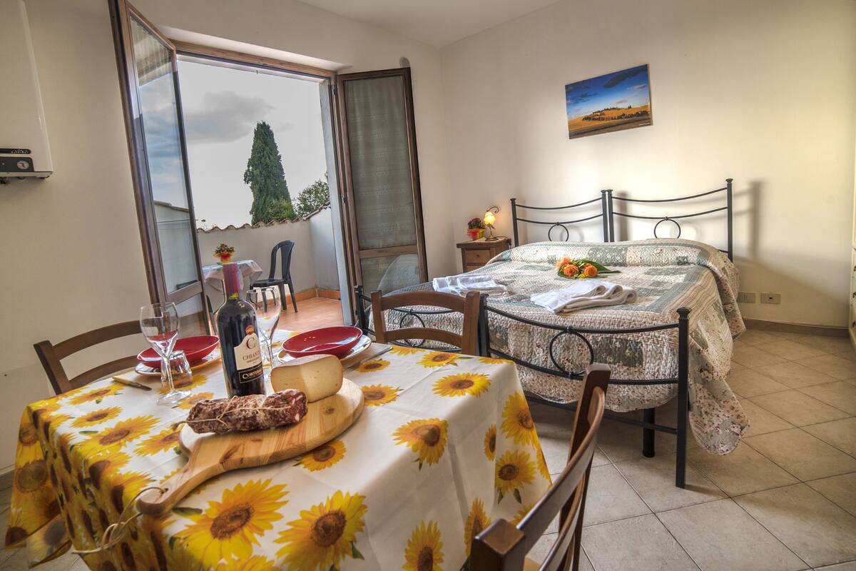 A dining area with a table set for two, featuring a wooden board with food and a bottle of wine. A bed with a floral bedspread is visible in the background.