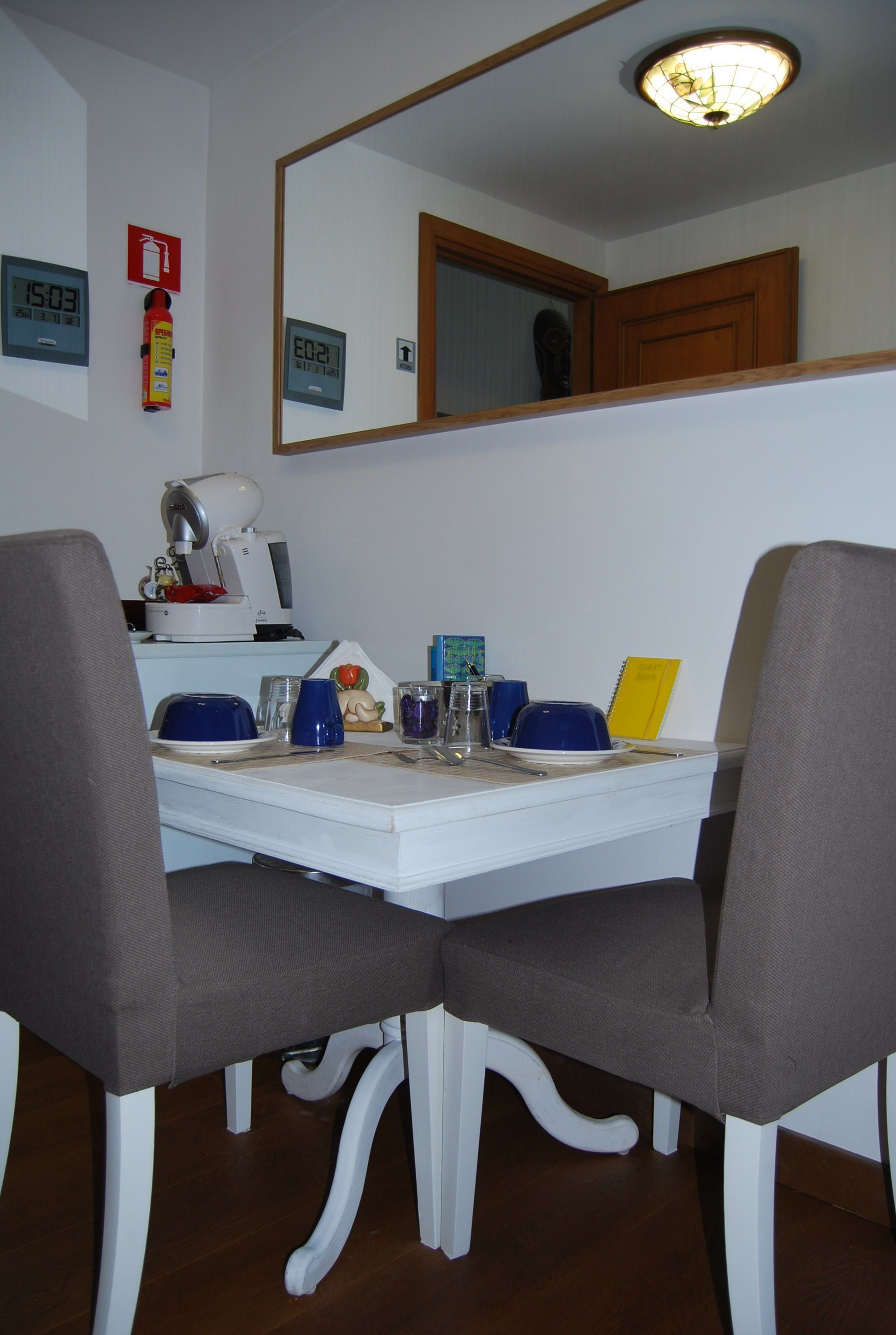 Uma sala de jantar com uma mesa branca, duas cadeiras cinzas, uma máquina de café e um extintor de incêndio na parede.