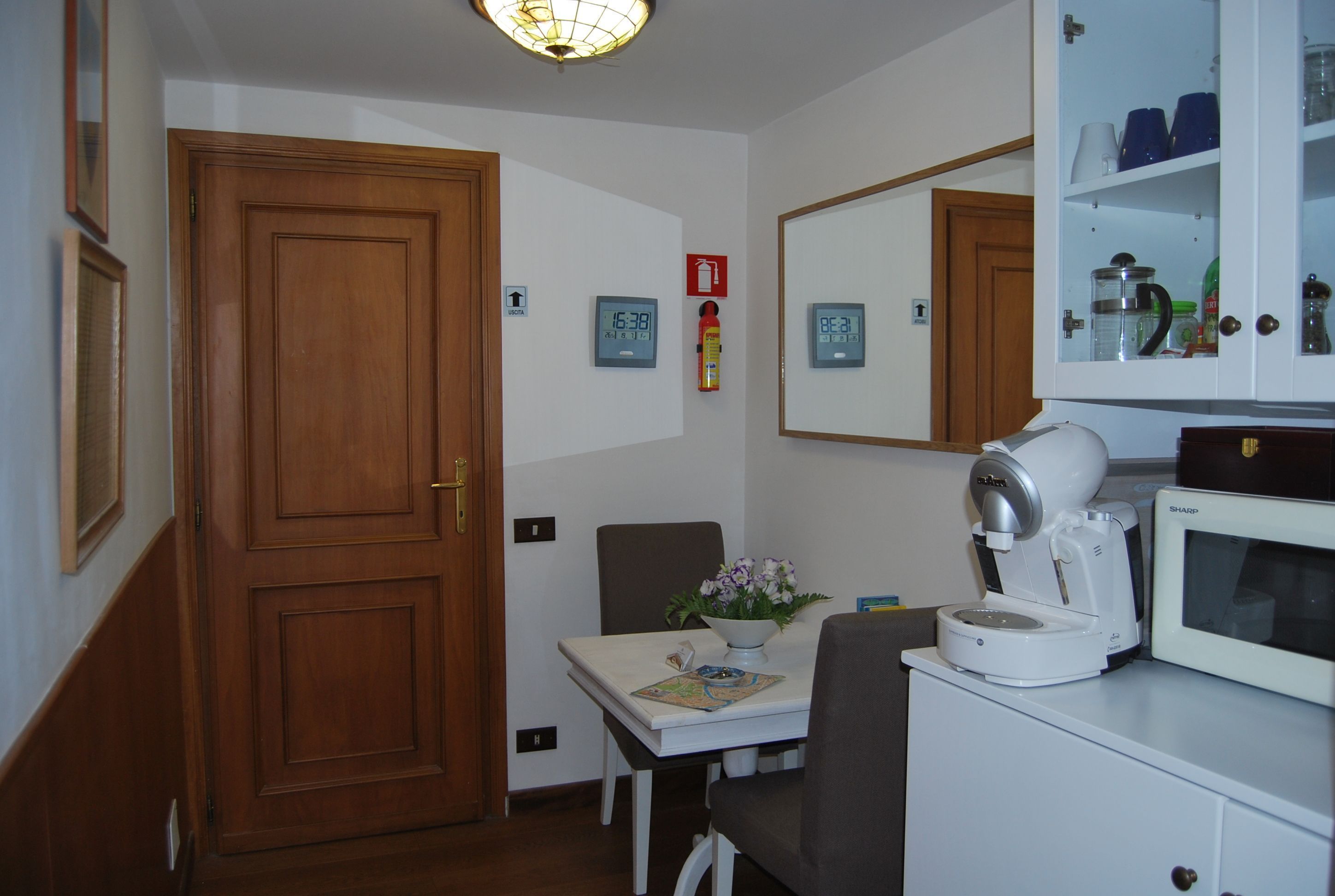 A small kitchen with a microwave, coffee maker, and cabinets. There is a table with a flower vase and a clock on the wall.