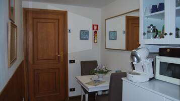 A small kitchen with a microwave, coffee maker, and cabinets. There is a table with a flower vase and a clock on the wall.