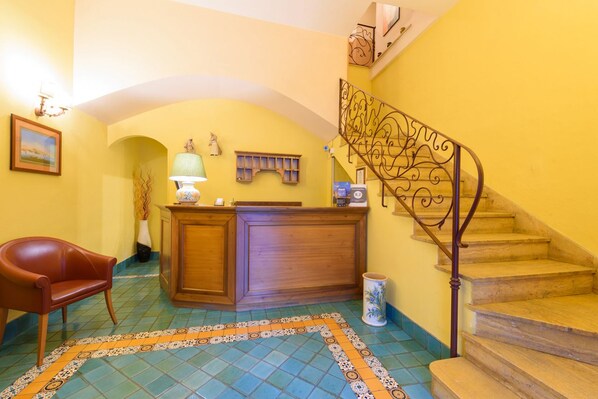A reception area with a wooden desk, a chair, a lamp, a decorative shelf, and a staircase with ornate railings. - Mergellina (Naples)