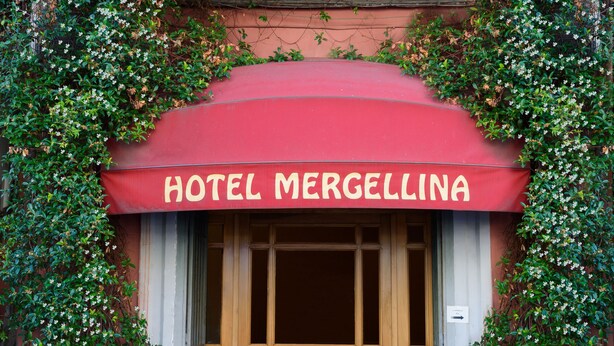 Ingresso di un hotel con tenda rossa, porte in legno e un balcone con persiane blu.