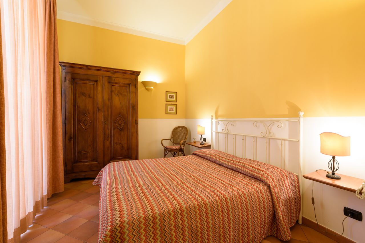 Une chambre à coucher comprenant un lit, une armoire en bois, une chaise, une table de chevet avec une lampe et un luminaire fixé au mur.