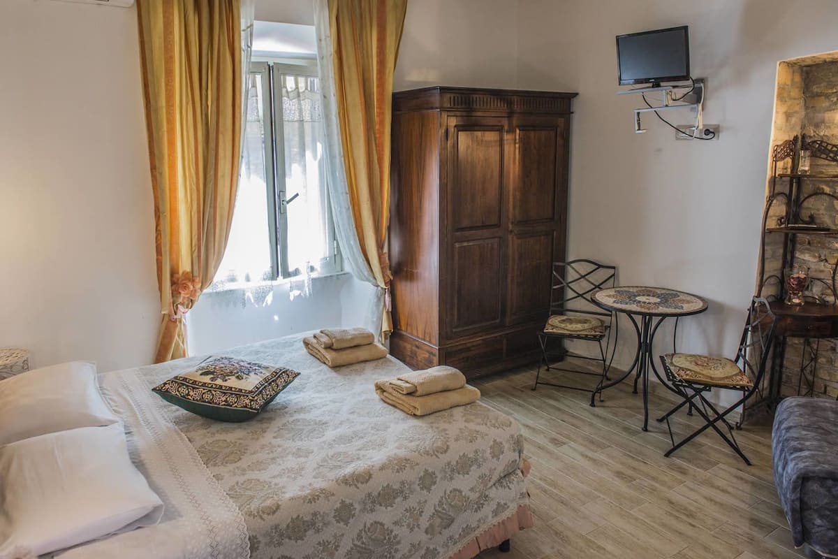 A hotel room with a bed, a wardrobe, a TV mounted on the wall, a small round table with chairs, and a window with curtains.