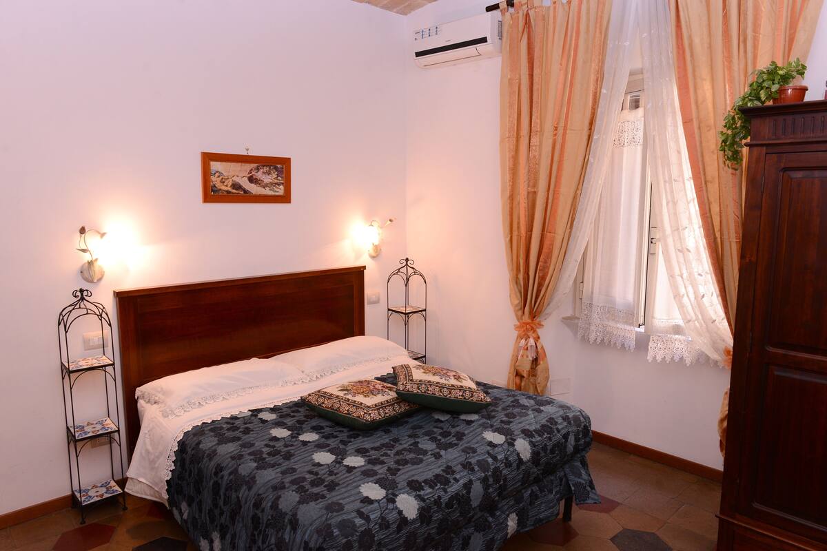 A bedroom with a wooden bed, two bedside tables with lamps, a wall-mounted air conditioner, and a window with sheer curtains.