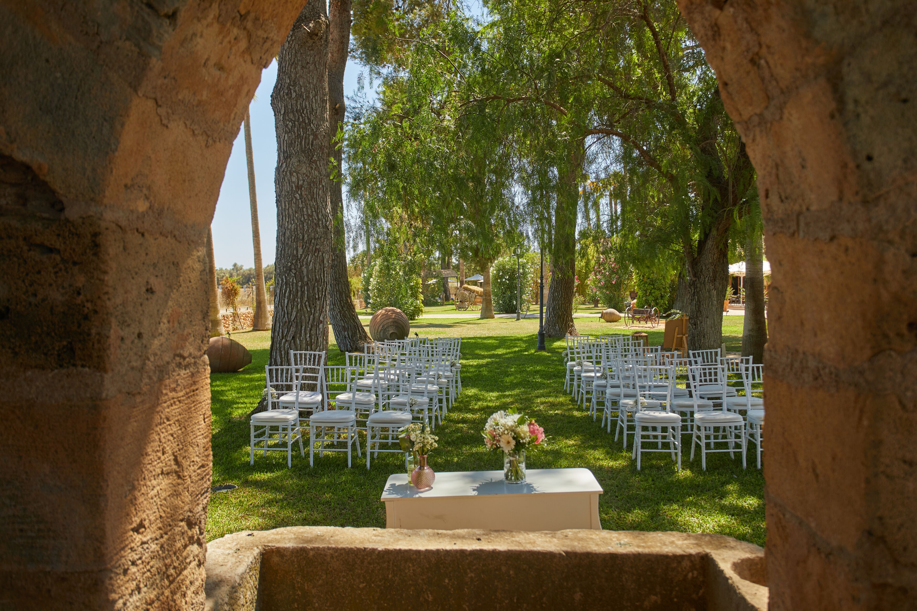 An outdoor wedding venue with white chairs arranged in rows, a central table with floral arrangements, and large trees providing shade.