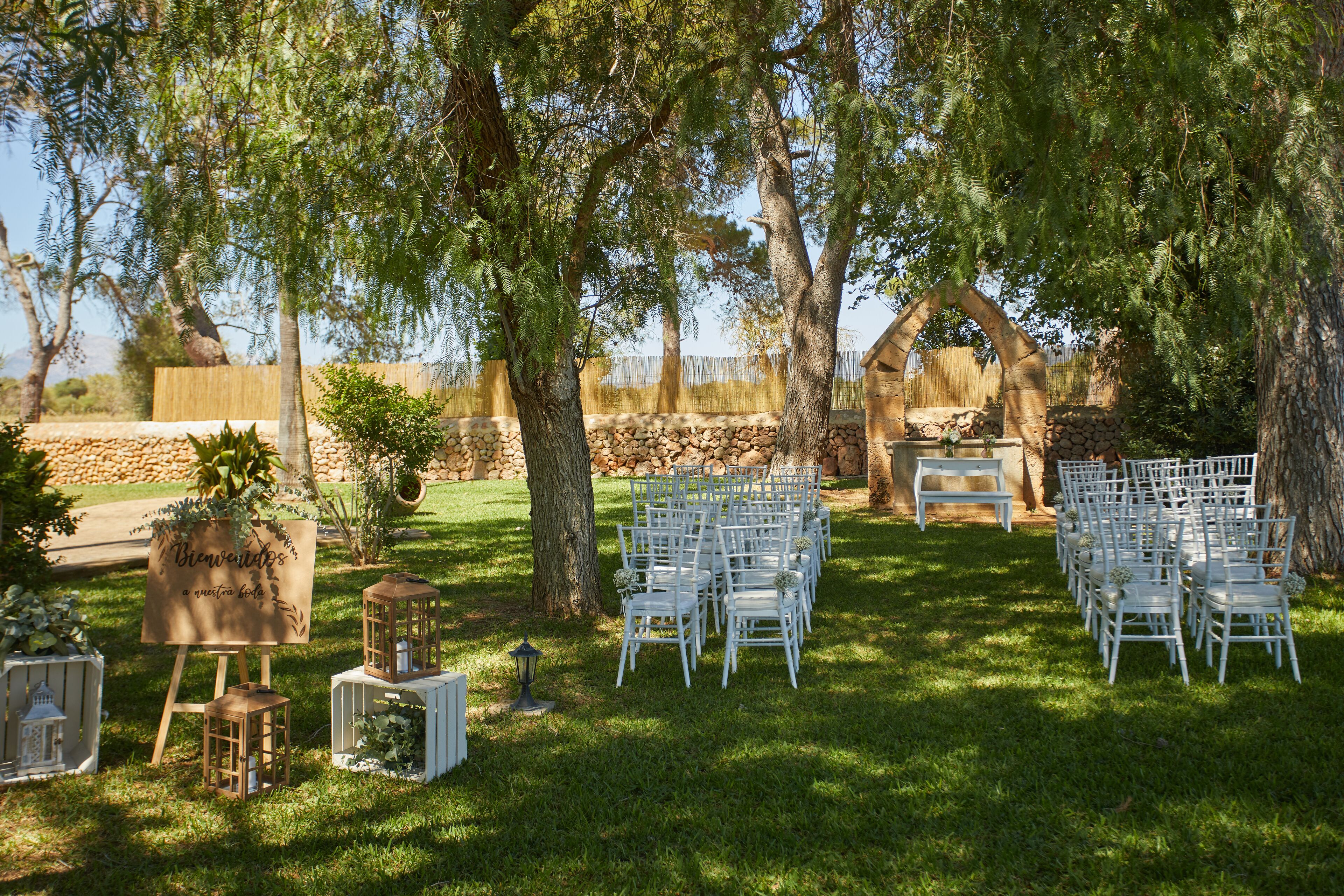 An outdoor wedding setup with white chairs arranged in rows, a wooden arch, and a sign that reads "Bienvenidos a nuestra casa."