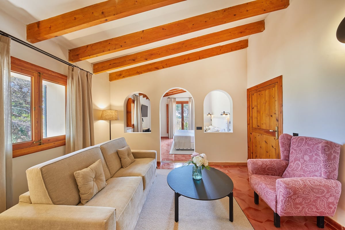 Un salon comprenant un canapé, un fauteuil et une table basse. Le plafond est en bois et, à travers une arche, on aperçoit une salle de bain.