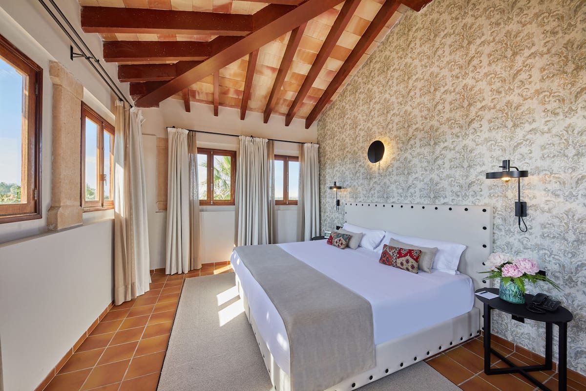 Une chambre à coucher avec un grand lit, des poutres apparentes en bois au plafond, un papier peint à motifs et une table de chevet ornée d’un vase de fleurs.