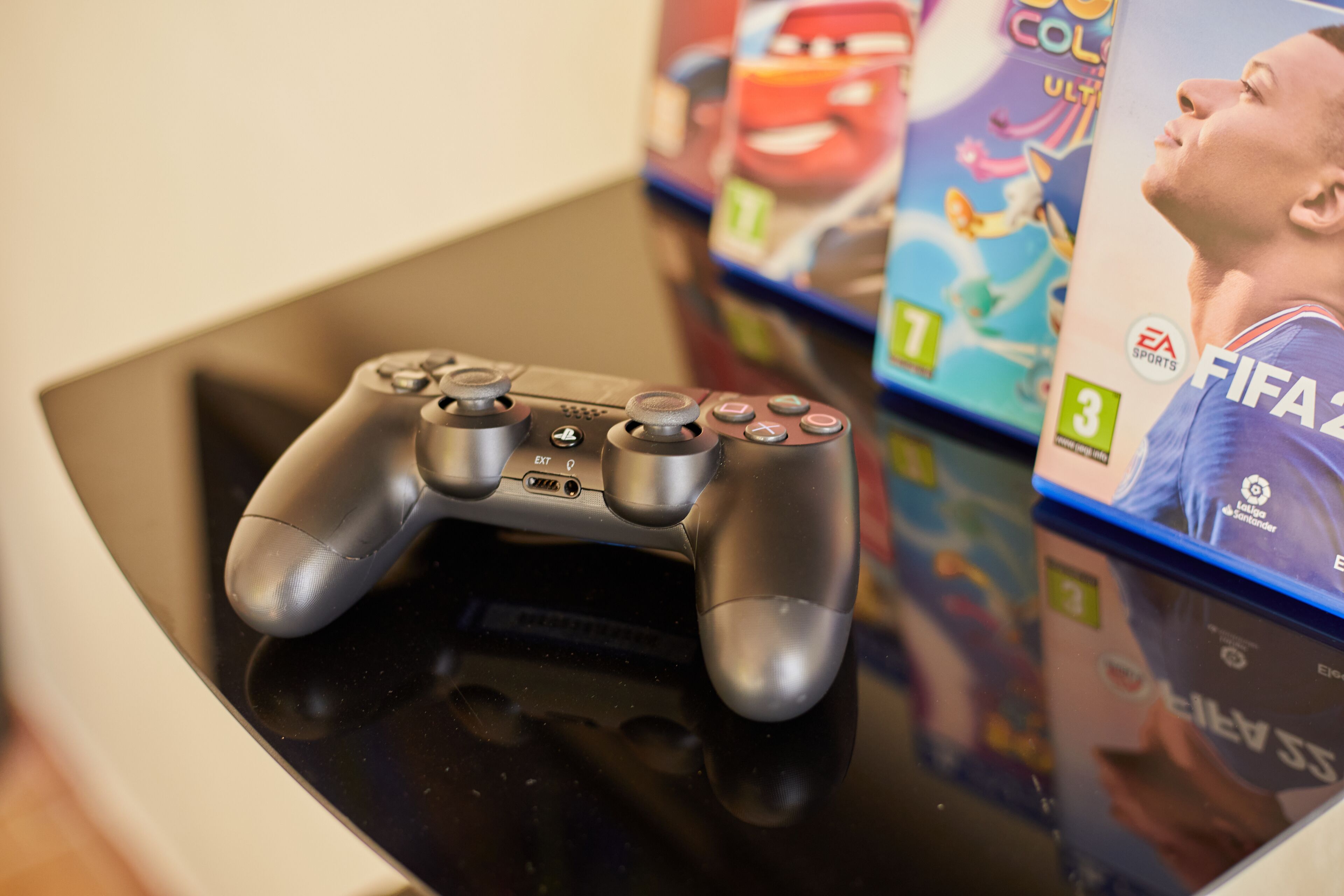 Un control de videojuego sobre una superficie reflectante, con fundas de videojuegos al fondo.