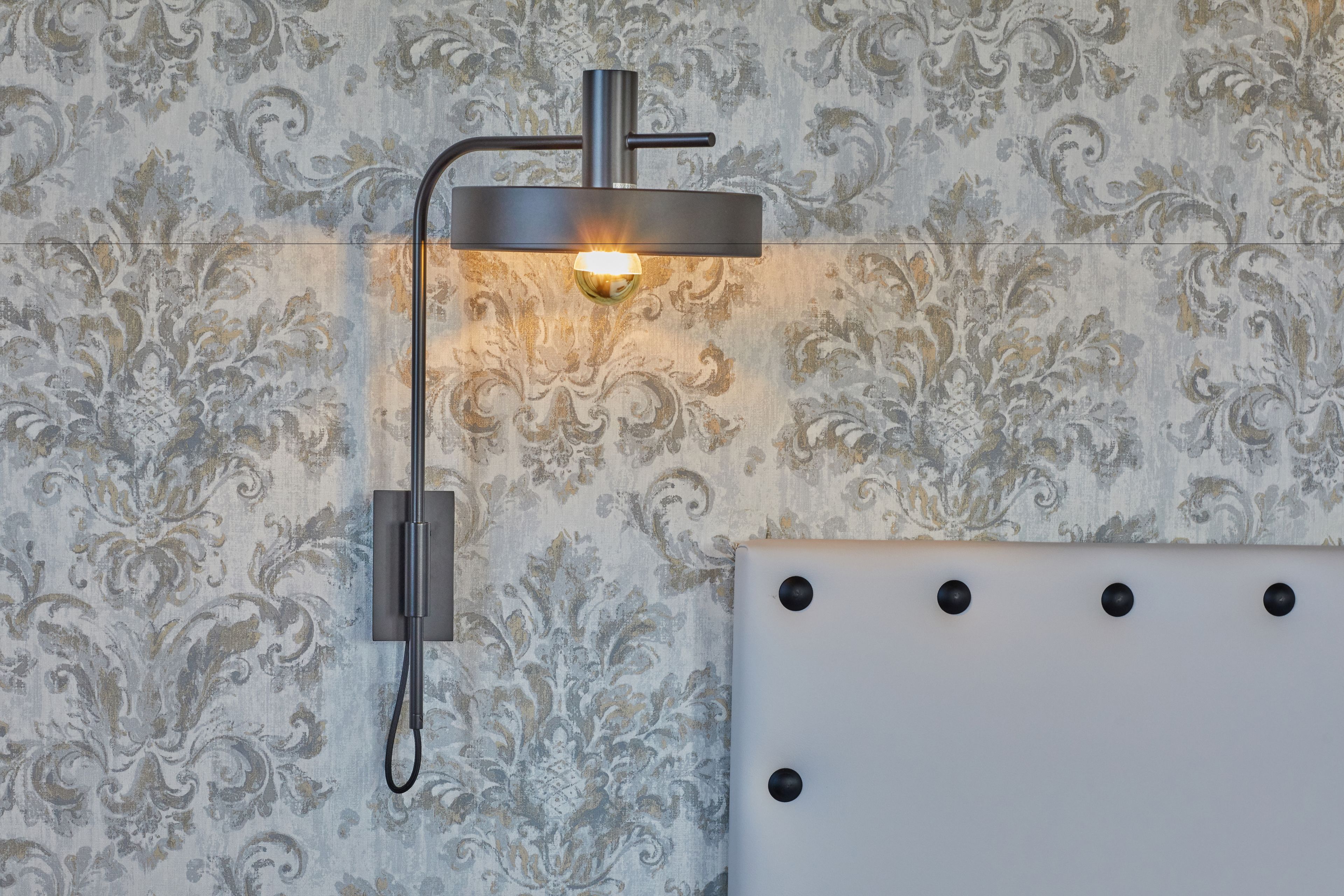A wall-mounted light fixture with a modern design, a textured wallpaper background, and a white panel with black rivets.
